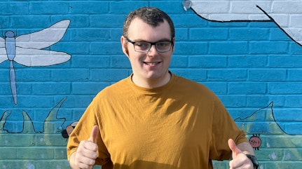 Image shows Mathew smiling and giving two thumbs up with a bright mural sky painted in the background