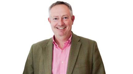 Image shows Michael smiling wearing a pink shirt and green jacket against a white background